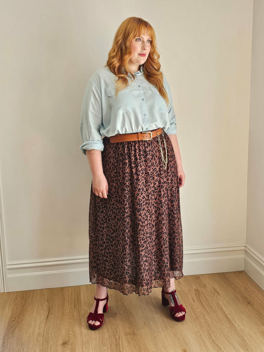 Woman wearing a light blue shirt, leopard print skirt, and red shoes against a plain wall.