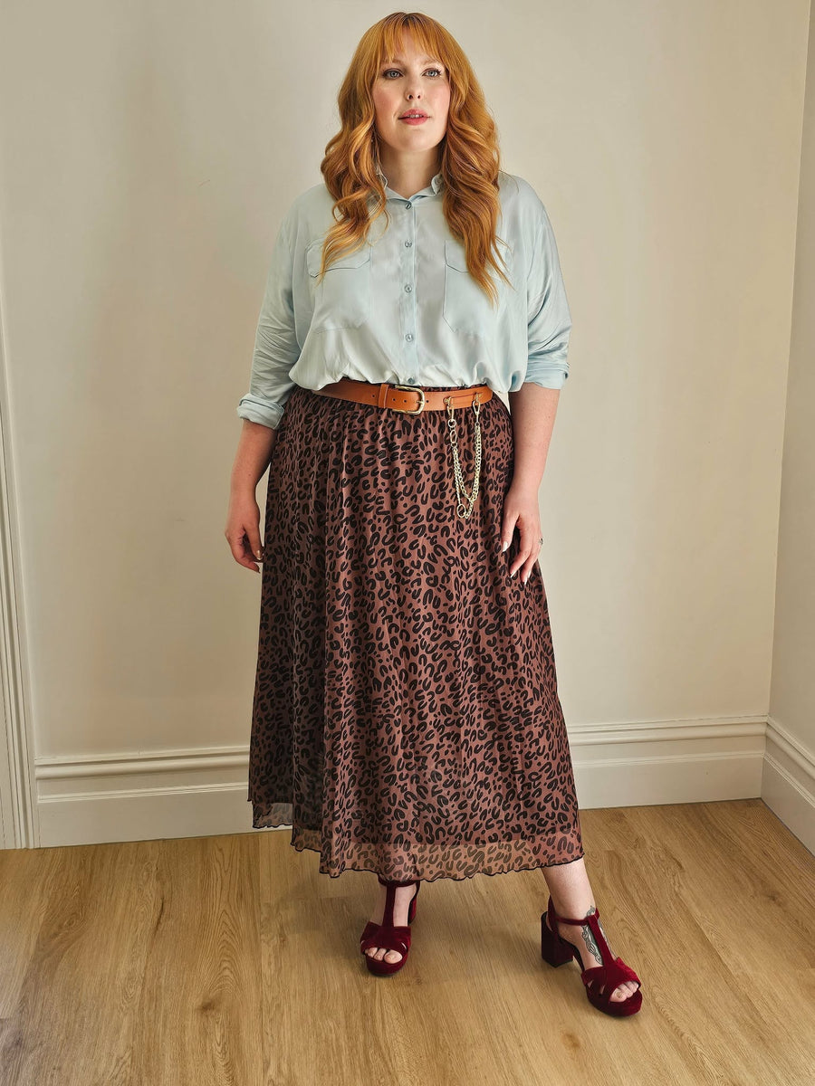 Woman wearing a light blue shirt, brown leopard print skirt, and red sandals against a beige wall.