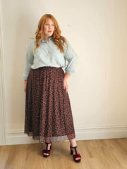 Woman wearing a light blue shirt and patterned skirt against a plain wall.