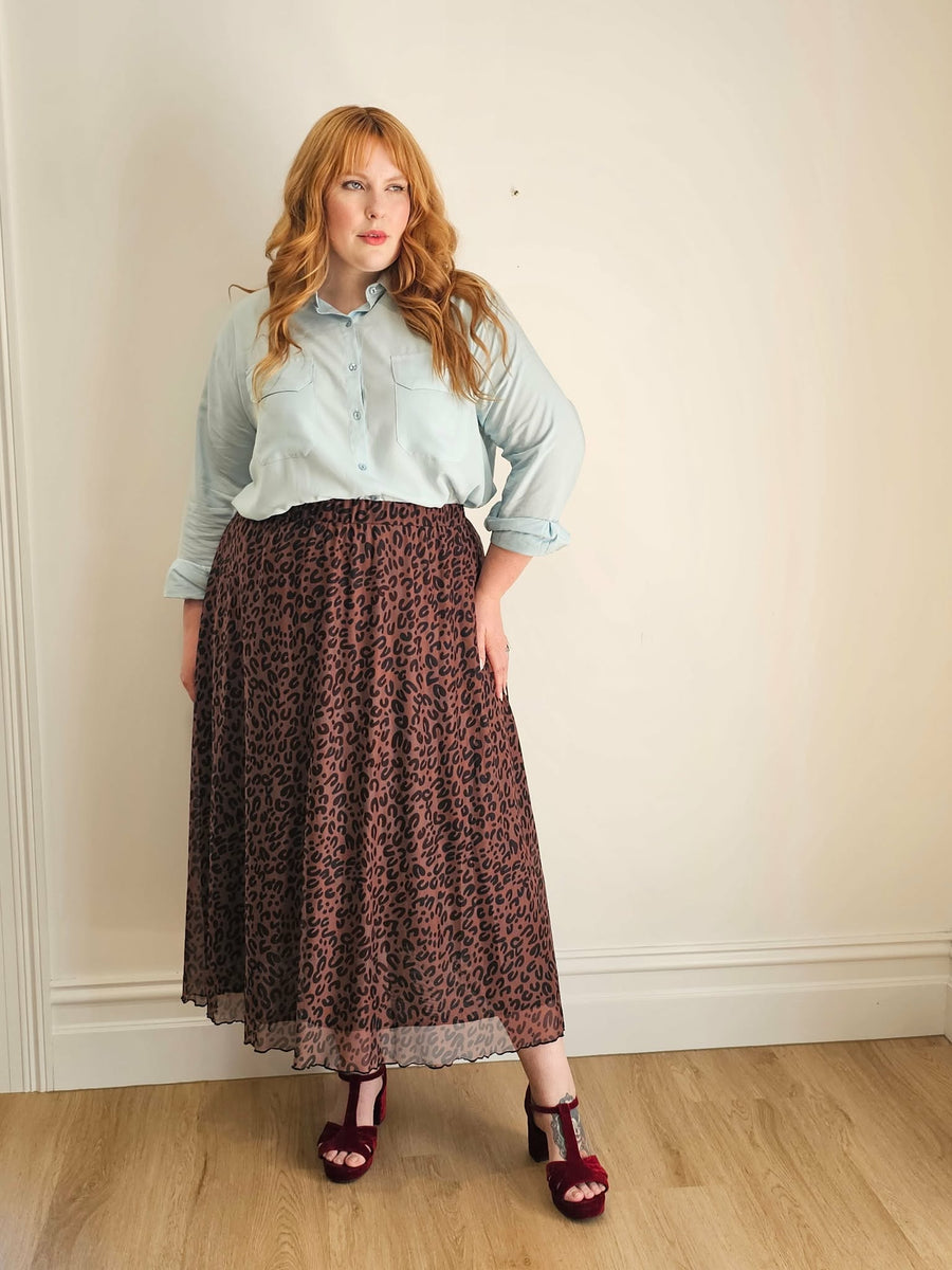 Woman wearing a light blue shirt and patterned skirt against a plain wall.