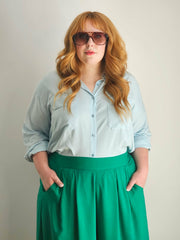 Woman wearing a light blue shirt and green skirt with sunglasses on a plain background