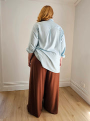 Person wearing a light blue shirt and brown pants standing in a room with white walls and wooden flooring.