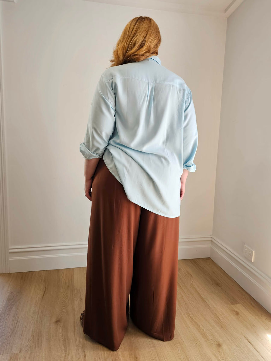Person wearing a light blue shirt and brown pants standing in a room with white walls and wooden flooring.